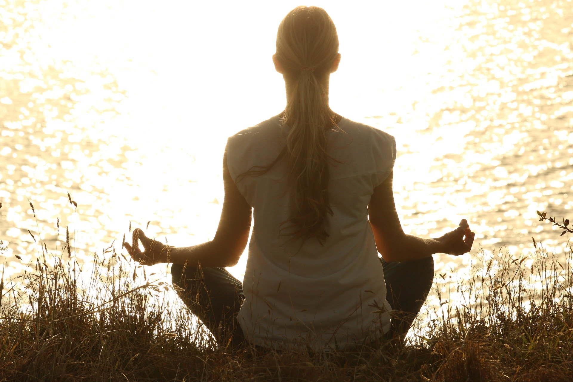 A woman meditating in the grass while facing the water as the sun is setting. 