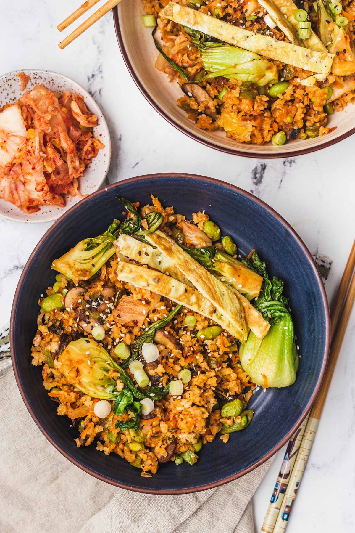 A birds-eye view of the kimchi fried rice on two plates, one blue plate and one beige plate, and a separate bowl of kimchi on the side topped with fried egg, and sesame seeds.