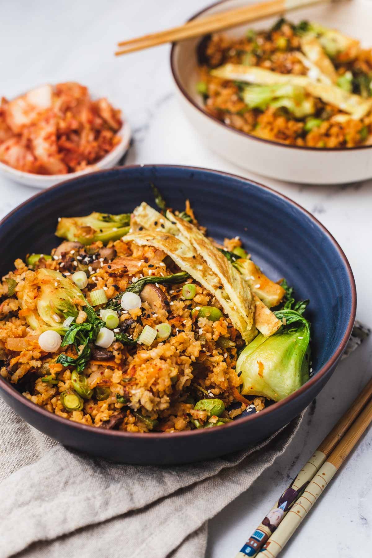 A close up of the fried rice dish in a blue bowl topped with green onions and vegetables with kimchi and a second bowl in the background.