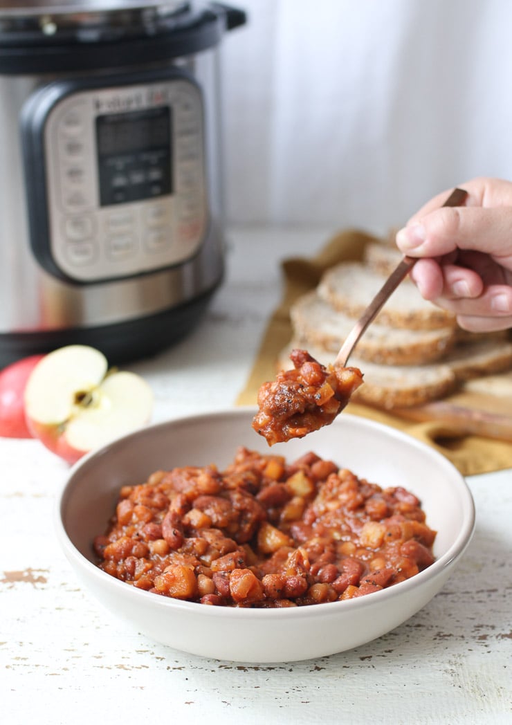 Instant pot baked beans in a white bowl. Someone is scooping a spoonful of baked beans. There is an instant pot, apples, and bread in the background. 