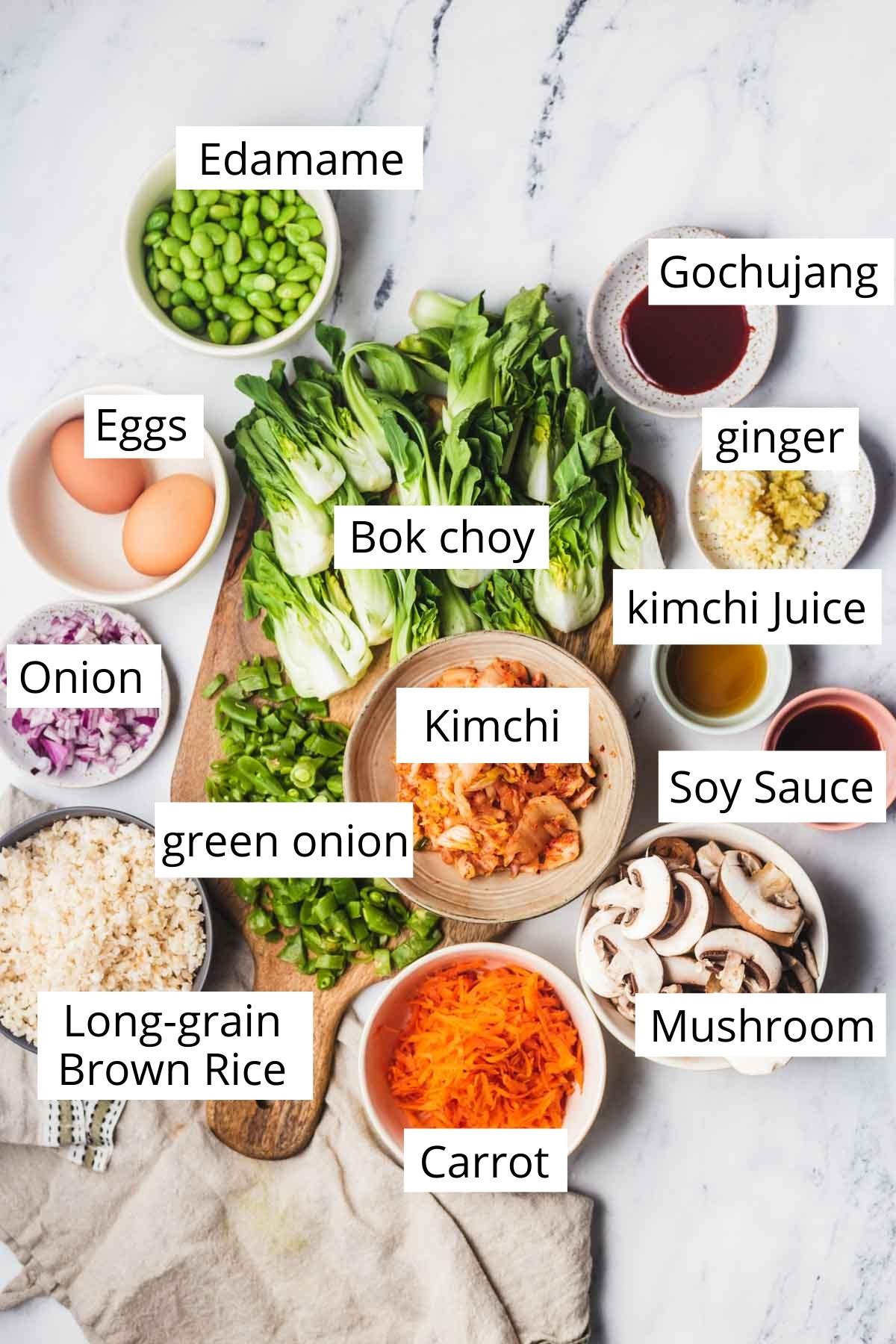 A birds-eye shot of the ingredients used for the kimchi fried rice recipe including gochujang, eggs, kimchi, kimchi juice, long-grain brown rice, and vegetables including carrot, bok choy, mushroom, onion, snap pea, green onion, garlic and edamame. 