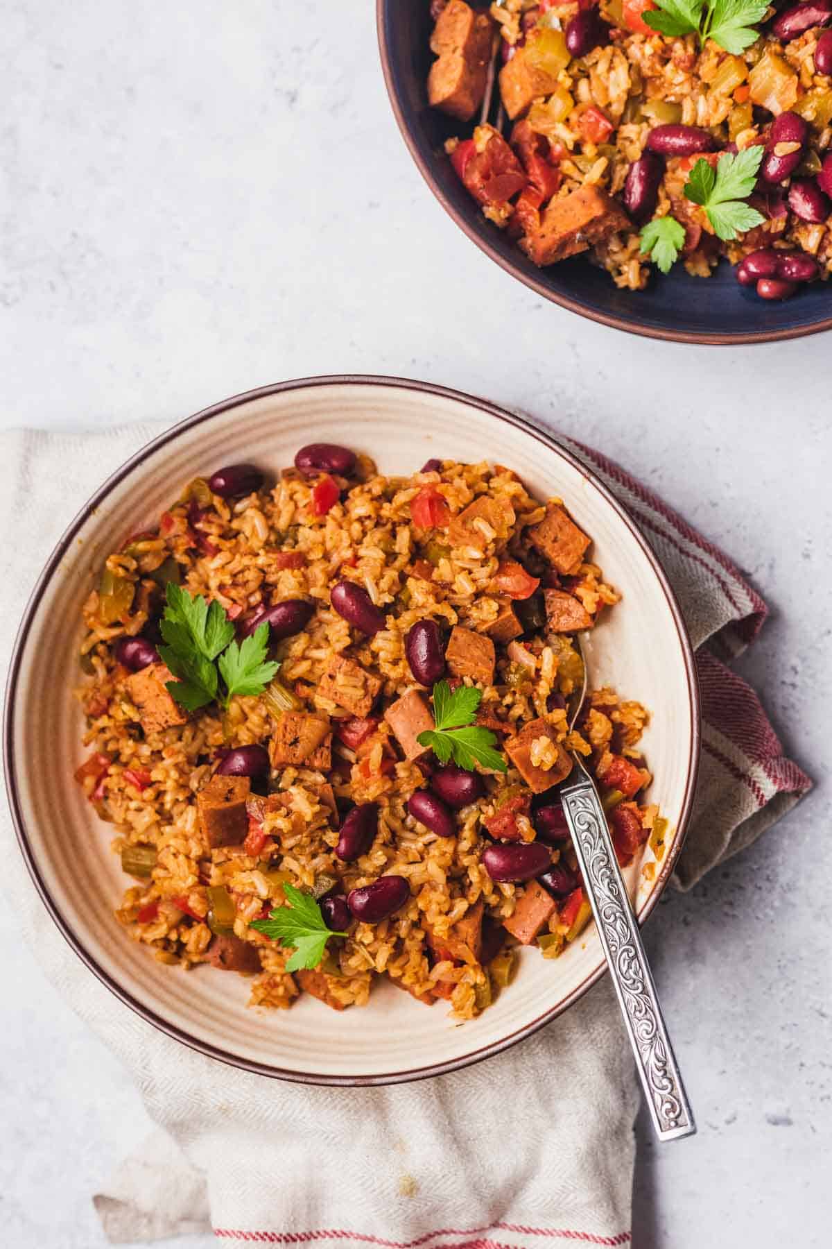 Birds eye view of a bowl of vegan jambalaya in a white bowl on top of a red and white striped dish towel. There is a quarter of another bowl of jambalaya in a blue bowl in the top right corner. 