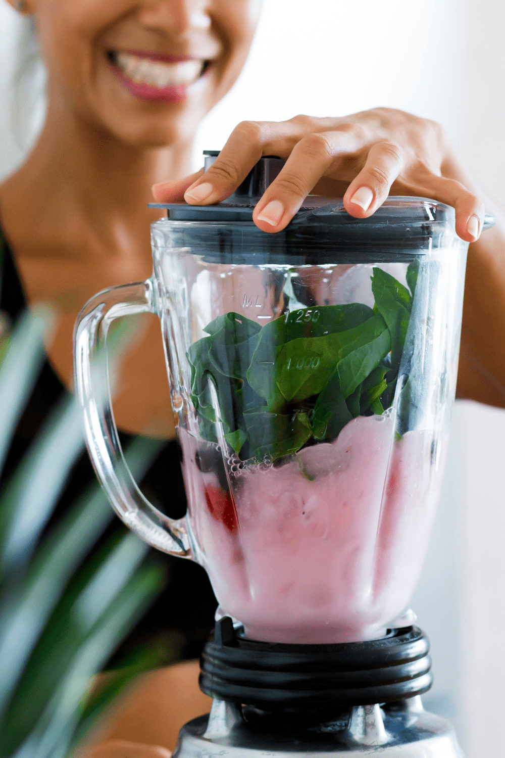 Close up of a blender with smoothie contents inside with a women smiling and holding the blender in the background.
