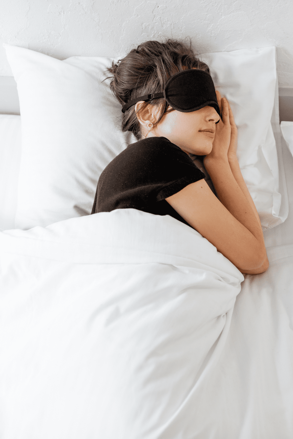Woman laying in bed on her side with a black eye mask on. 