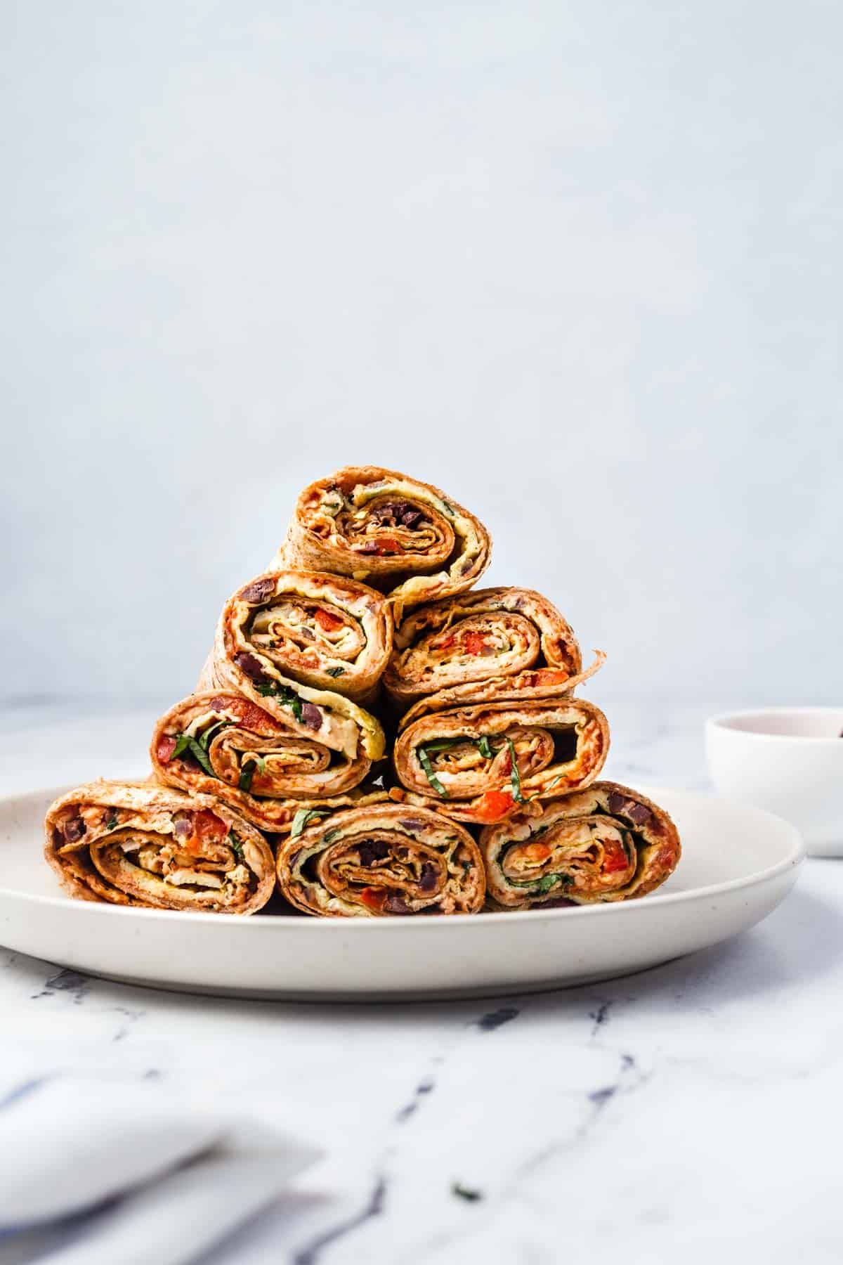 8 breakfast pizza rolls stacked on a white plate with a white bowl in the background.