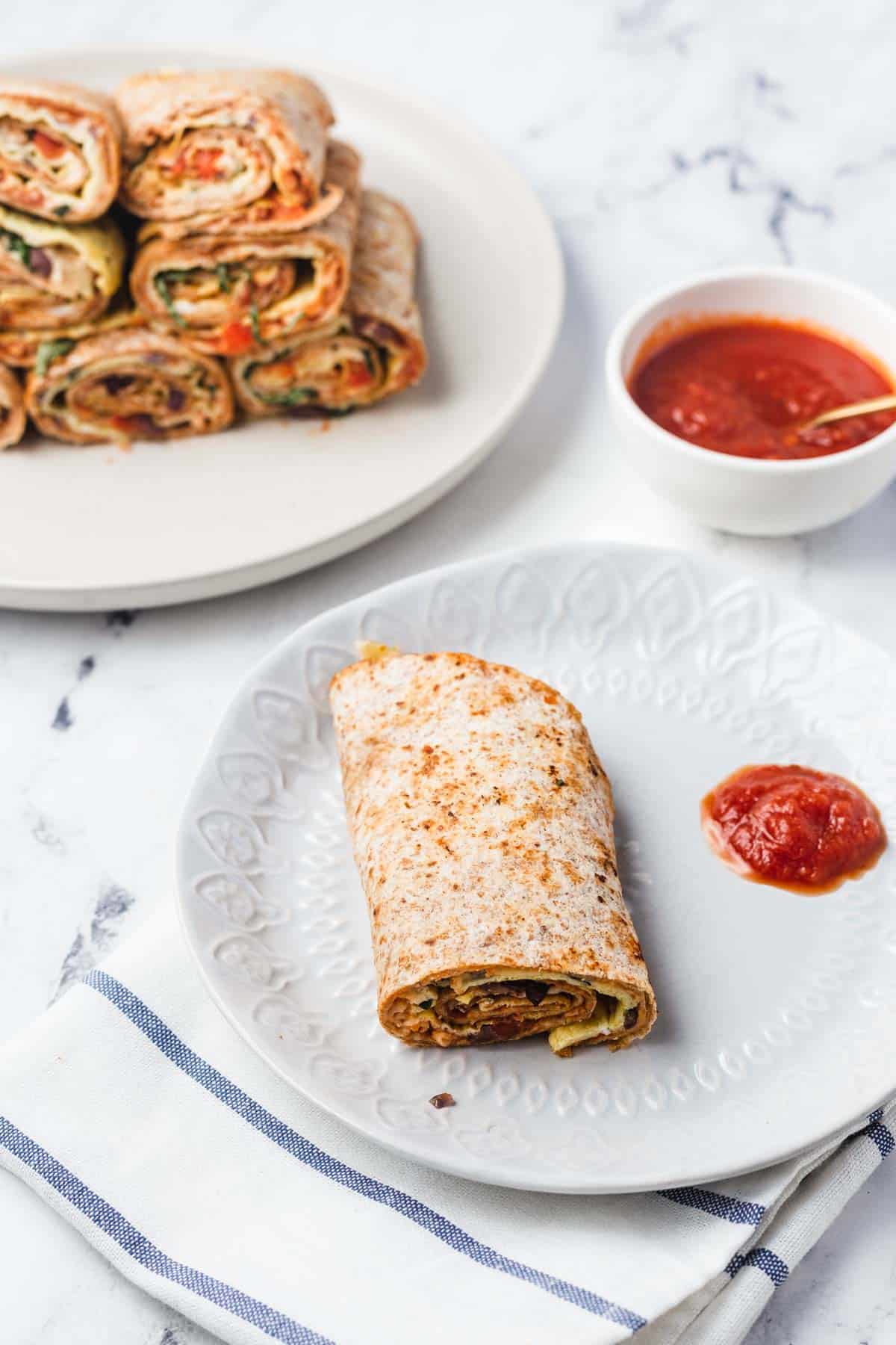 Birds eye view of a pizza roll on a white plate with pizza sauce on the side. There is another white plate with 7 pizza rolls stacked and a bowl of pizza sauce to the right of it. 