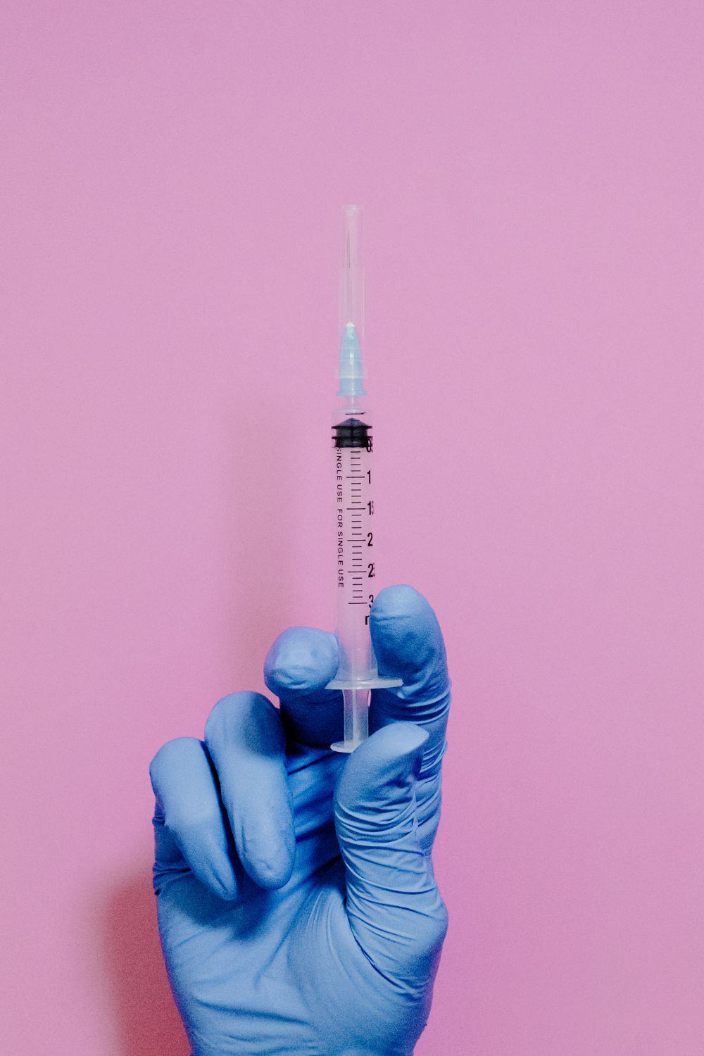 Picture of a hand with a blue latex glove holding an injection with a pink background.