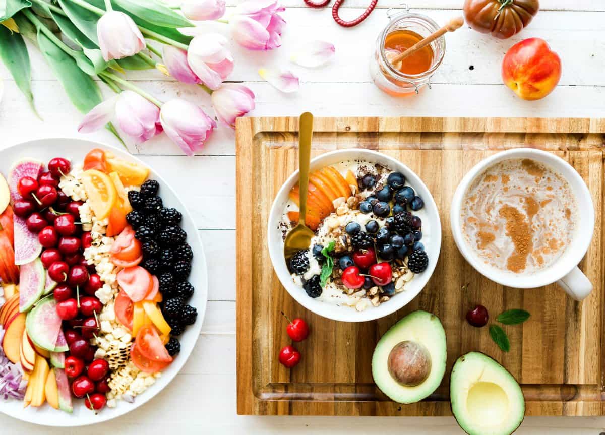 Birds eye view of a breakfast layout including fresh fruits, a yogurt bowl, and a latte on a wooden cutting board.