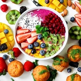 Purple smoothie bowl topped with various fruits, surrounded by an arrangement of colourful fruit.