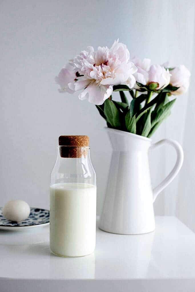 Clear glass of milk on a table with a vase of flowers behind it.