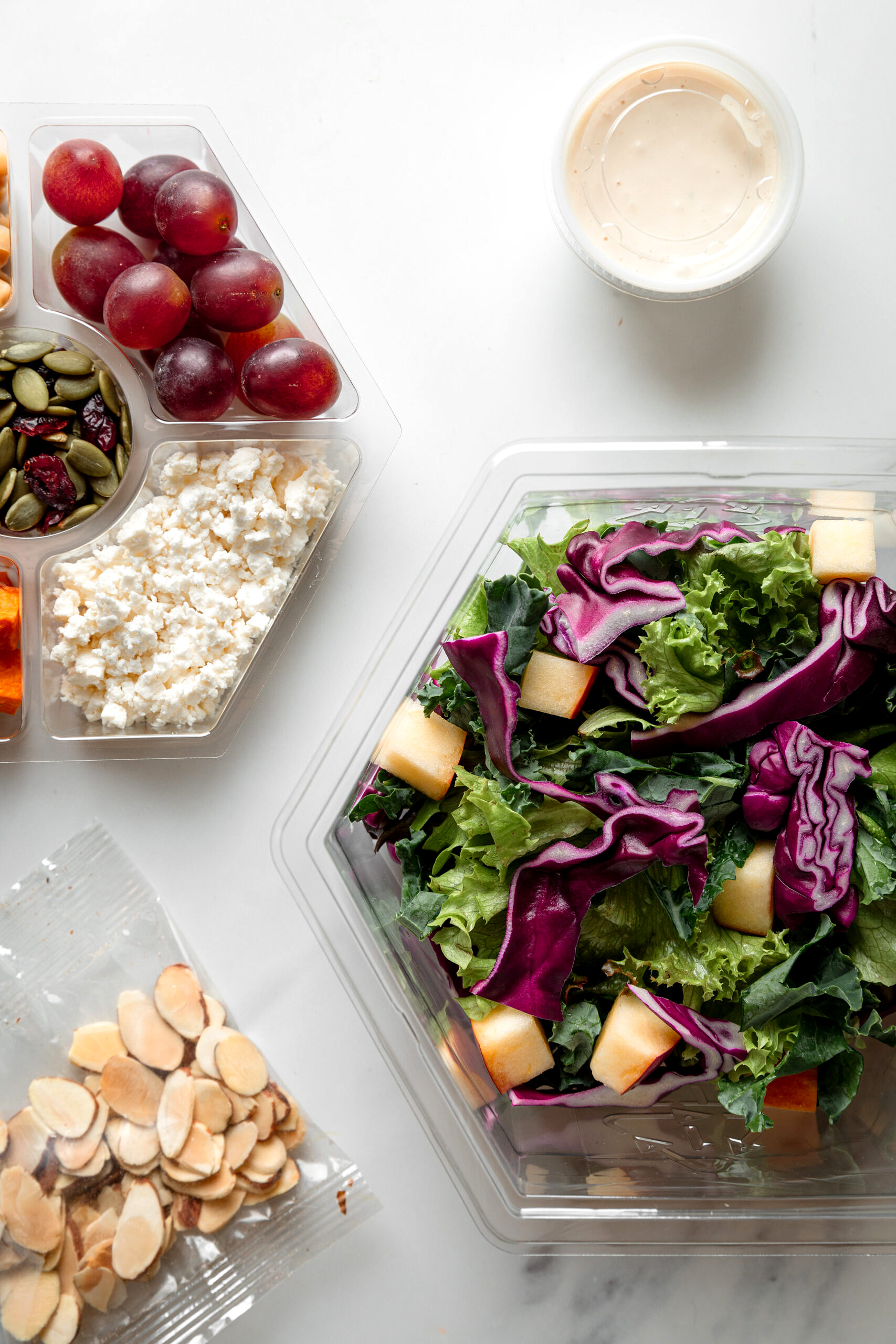 Three layers of tahini crunch salad, one with cabbage and kale base, one with feta, seeds and grapes, and a bag of almonds.