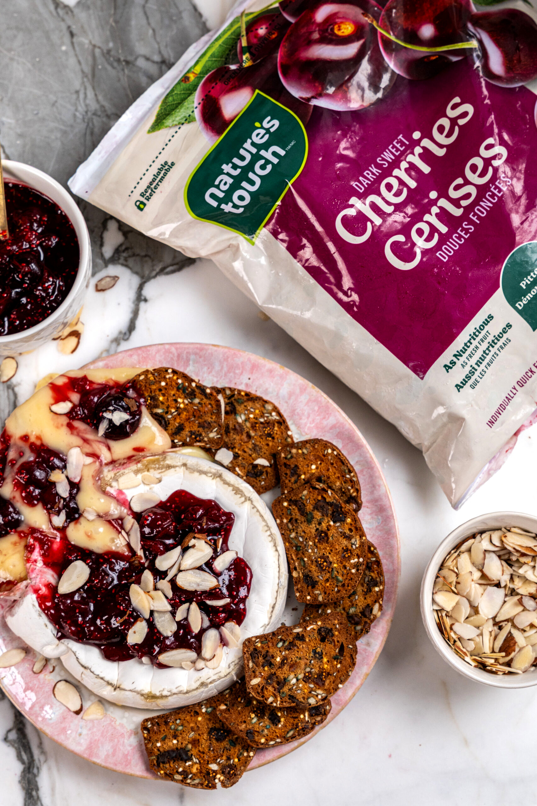 A plate of cherry chia baked brie served with crackers and sliced almonds, next to a bag of Nature's Touch frozen dark sweet cherries.
