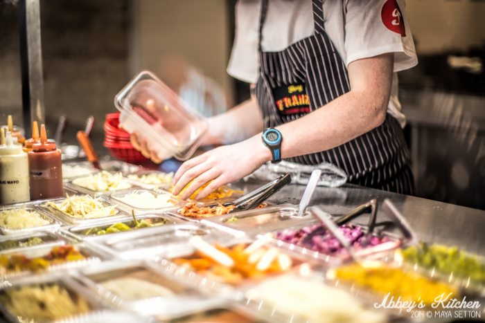 A person putting together the hot dog with ingredients in containers.